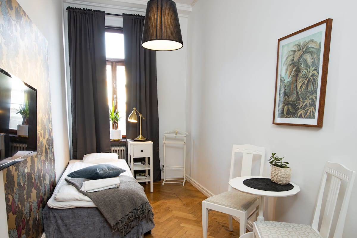 A bedroom with a bed, bedside table, lamp, chair, and a painting on the wall.