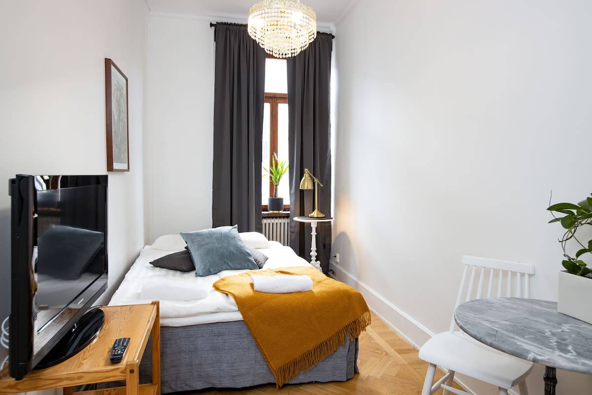 A hotel room with a bed, a TV, a bedside table with a remote, a small table with a chair, and a window with curtains.