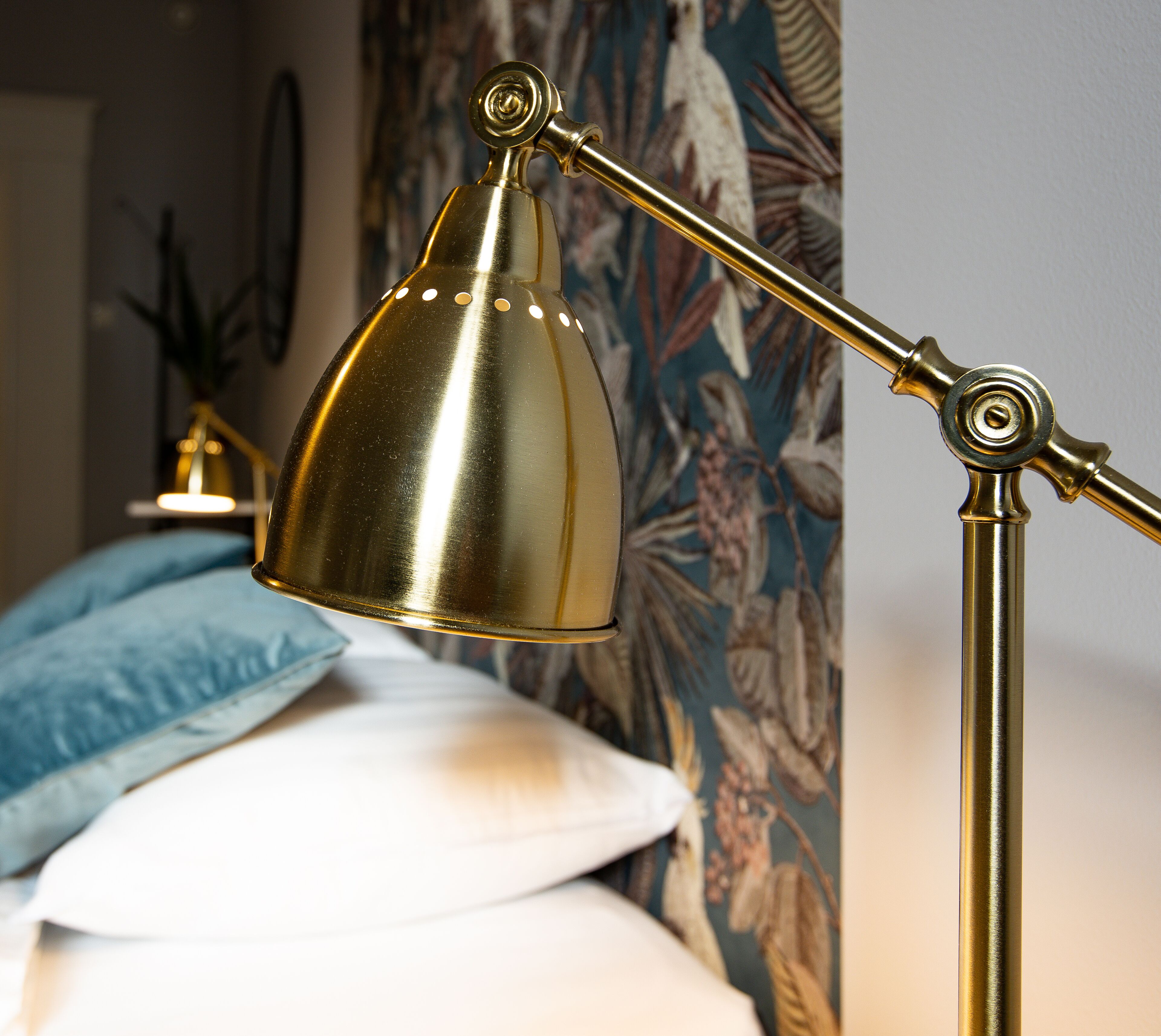 A brass desk lamp with a conical shade, positioned in front of a bed with a floral-patterned headboard.