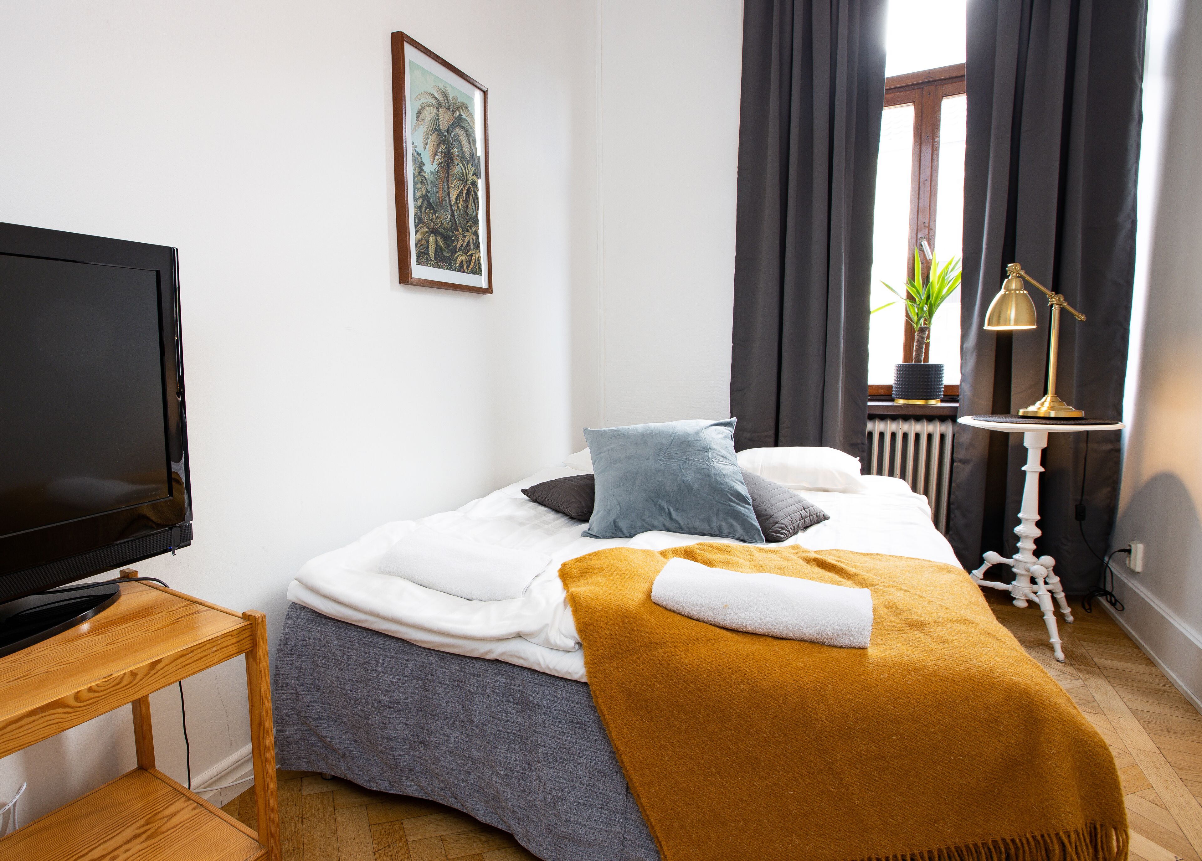 A neatly made bed with a white comforter, a mustard-colored throw blanket, and a single pillow. A television is placed on a wooden stand to the left.