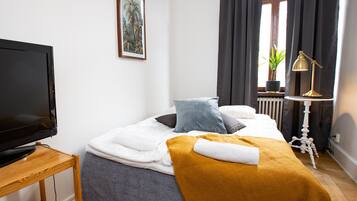 Un lit bien fait, recouvert d’une couette blanche, d’un plaid de couleur moutarde et d’un seul oreiller. Un téléviseur est posé sur un meuble en bois, à gauche.