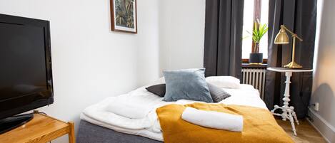 A neatly made bed with a white comforter, a mustard-colored throw blanket, and a single pillow. A television is placed on a wooden stand to the left.