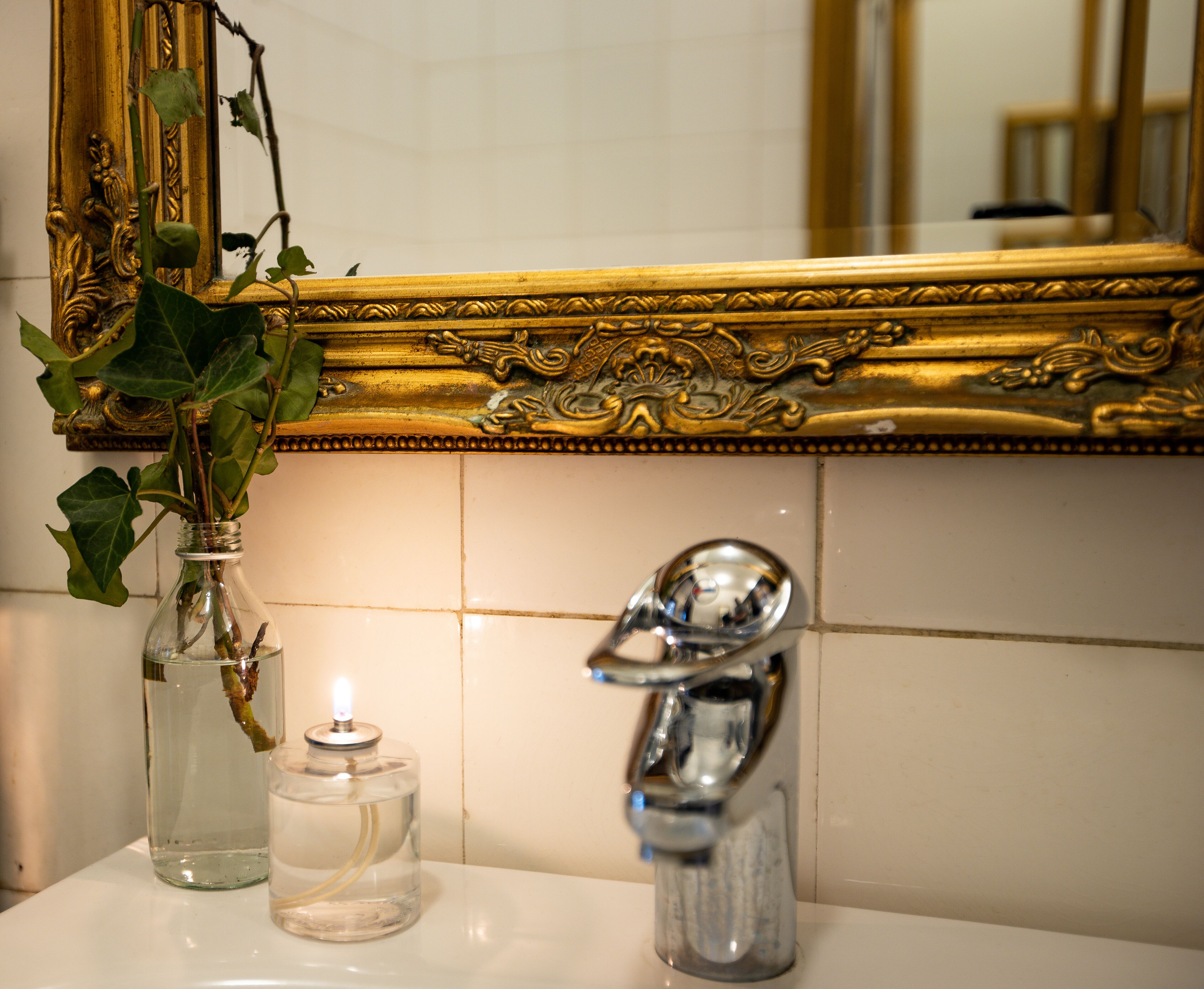 A bathroom with a gold-framed mirror, a glass vase with a plant, and a clear glass candle holder.