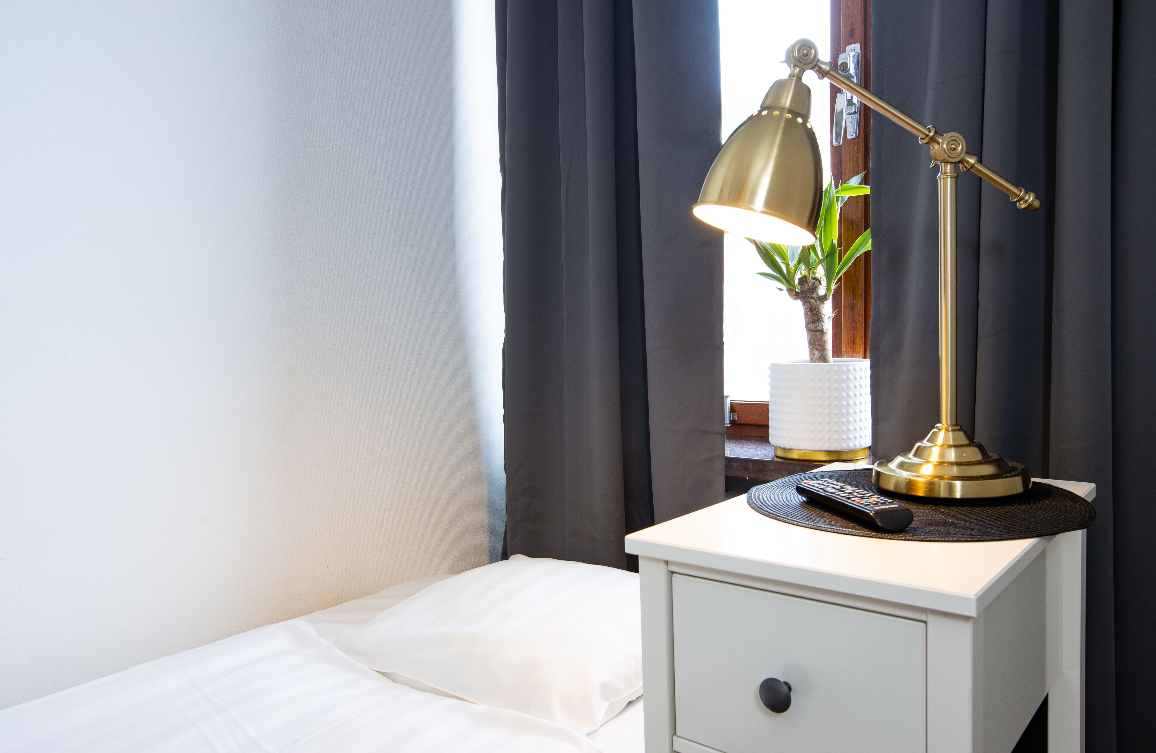 En pænt redt seng med hvide sengetøj, et natbord med en lampe, en fjernbetjening og en lille potteplante.
