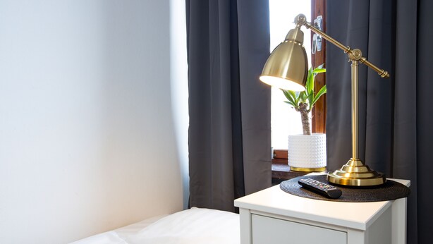 A neatly made bed with white linens, a bedside table with a lamp, a remote control, and a small potted plant.
