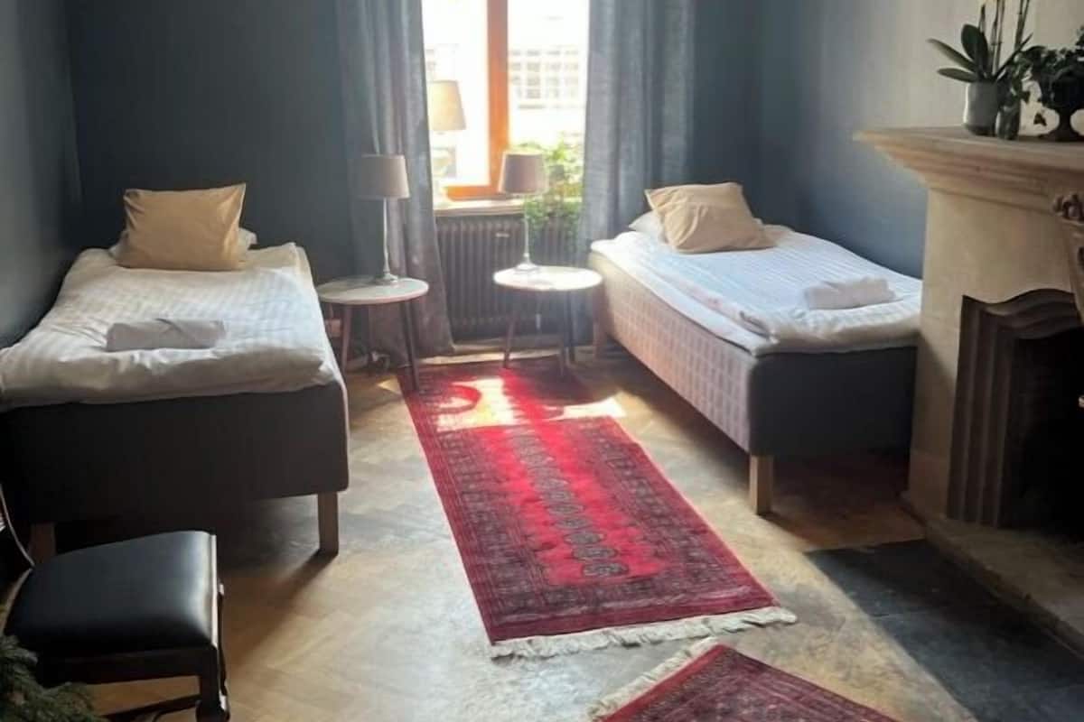 A compact hotel room with two single beds, a small round table, a chair, a fireplace, and a large window with curtains.