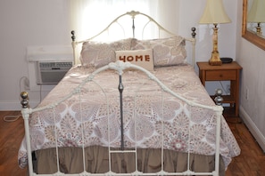 Un lit avec un cadre en métal, une literie à motifs, un coussin orné du mot «HOME», une lampe de chevet et une petite table de nuit en bois.