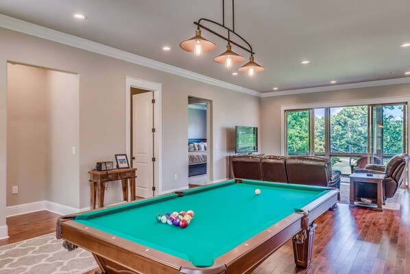 A pool table with balls, a sofa, and a television in a room with large windows.