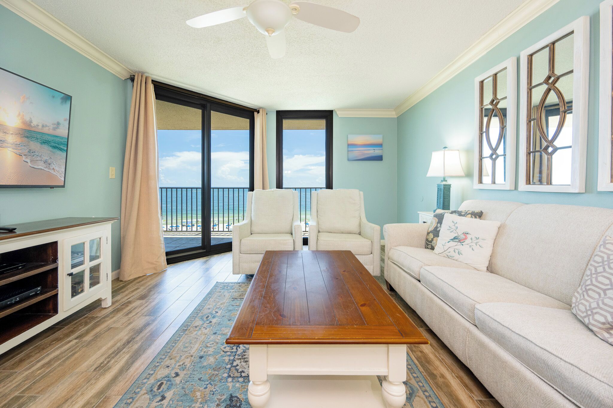 A living room with a wooden coffee table, two white armchairs, a beige sofa, a TV, and a view of the ocean through a sliding glass door.