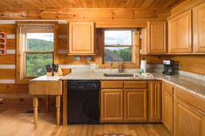 Une cuisine rustique, située dans une cabane en bois, équipée d’un lave-vaisselle, d’un évier et de fenêtres donnant sur une forêt.