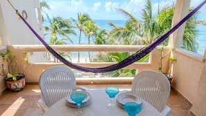 Een balkon met een hangmat, twee stoelen, een tafel en uitzicht op de oceaan en palmbomen.