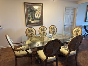 Un coin repas avec une table en verre entourée de chaises ornées de motifs de palmiers, un tableau encadré au-dessus, et une porte visible au fond.