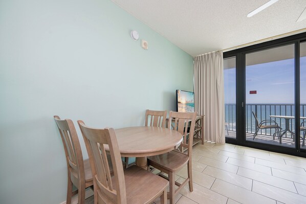 A room with a wooden dining table and chairs, a television, and a sliding glass door leading to a balcony with outdoor furniture.