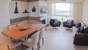 A modern living room with a wooden dining table, black chairs, a black sofa, and a large window offering a city view. - Gramado me Aptos de Luxo Via Florida (Gramado)