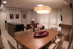 Uma área de refeições moderna com uma mesa de madeira, cadeiras bege, um centro de mesa decorativo vermelho, um sofá marrom com almofadas estampadas e iluminação pendente.