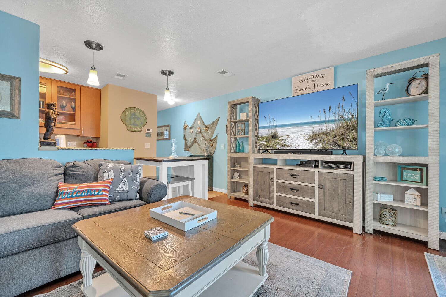 A living room with a grey sofa, wooden coffee table, and a TV on a wooden stand. There is a 'Welcome to our Beach House' sign above the TV.