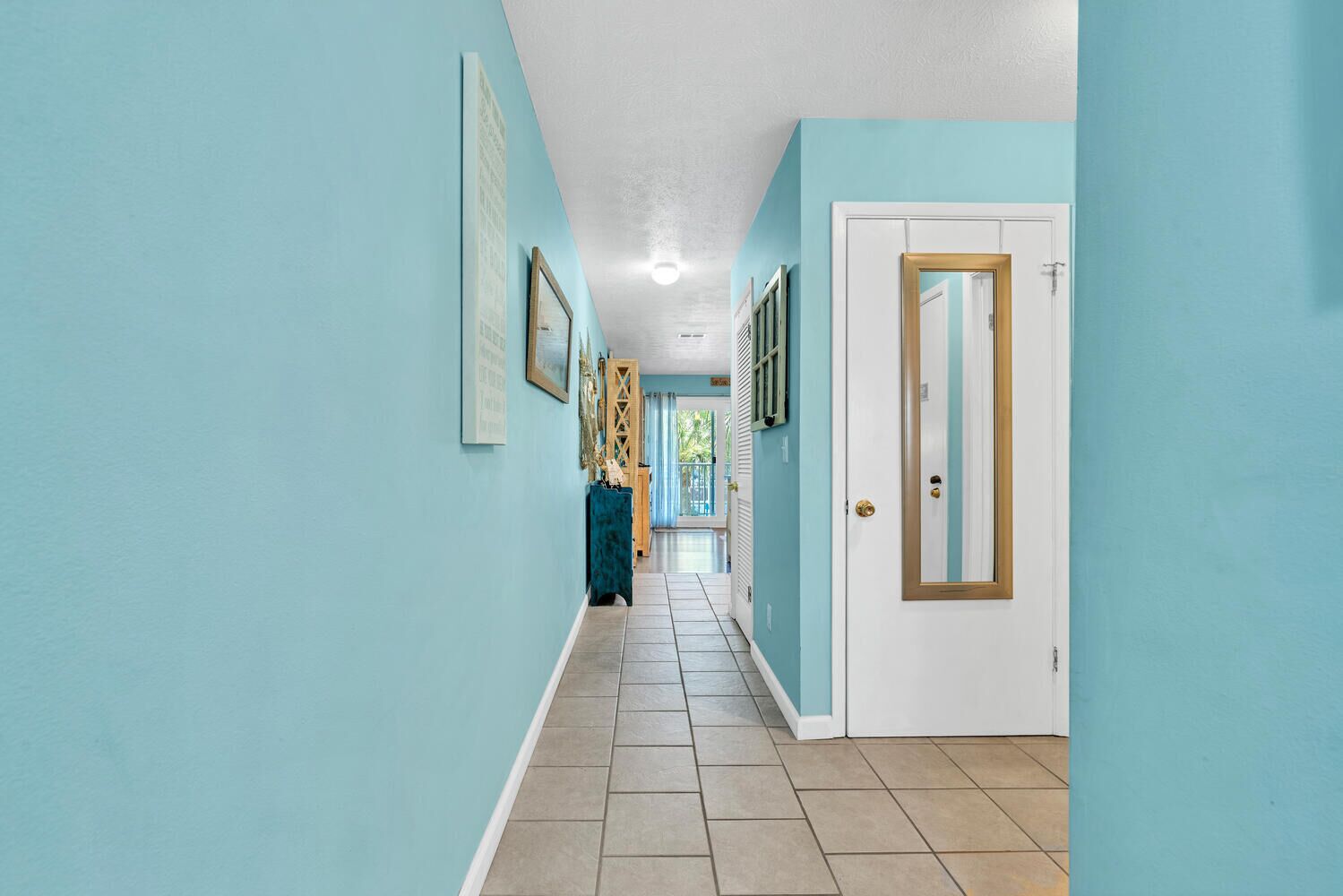 A hallway with light blue walls, a white door with a gold frame, and a tiled floor.
