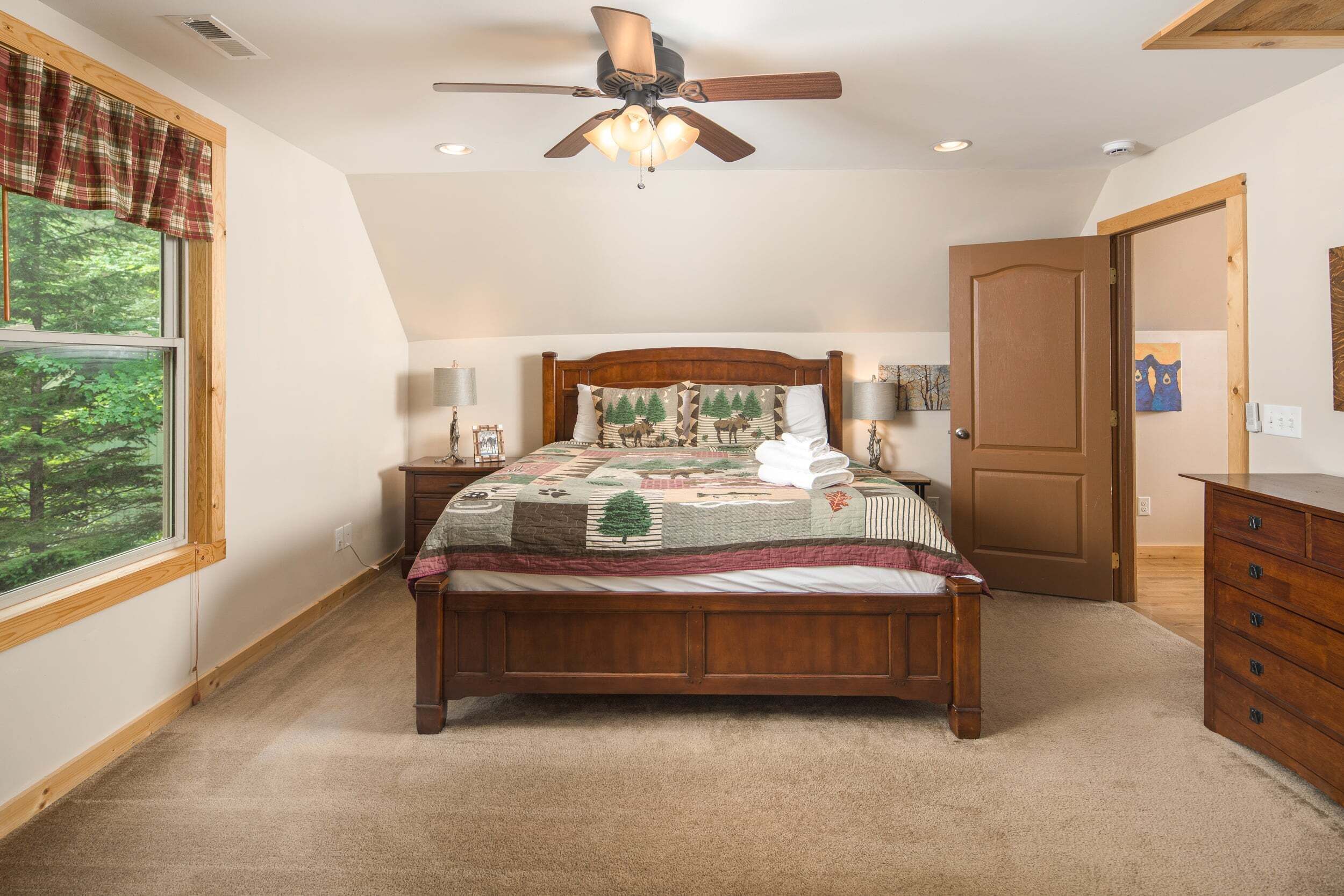 A bedroom with a wooden bed, a ceiling fan, a window with a view of trees, a door, and a dresser.