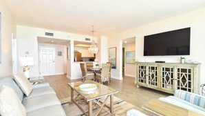 A modern living room with a glass coffee table, striped sofas, a dining area with wicker chairs, and a TV mounted on the wall. - Calm, cool, coastal beachfront condo w/private balcony! 🐚 (Destin)