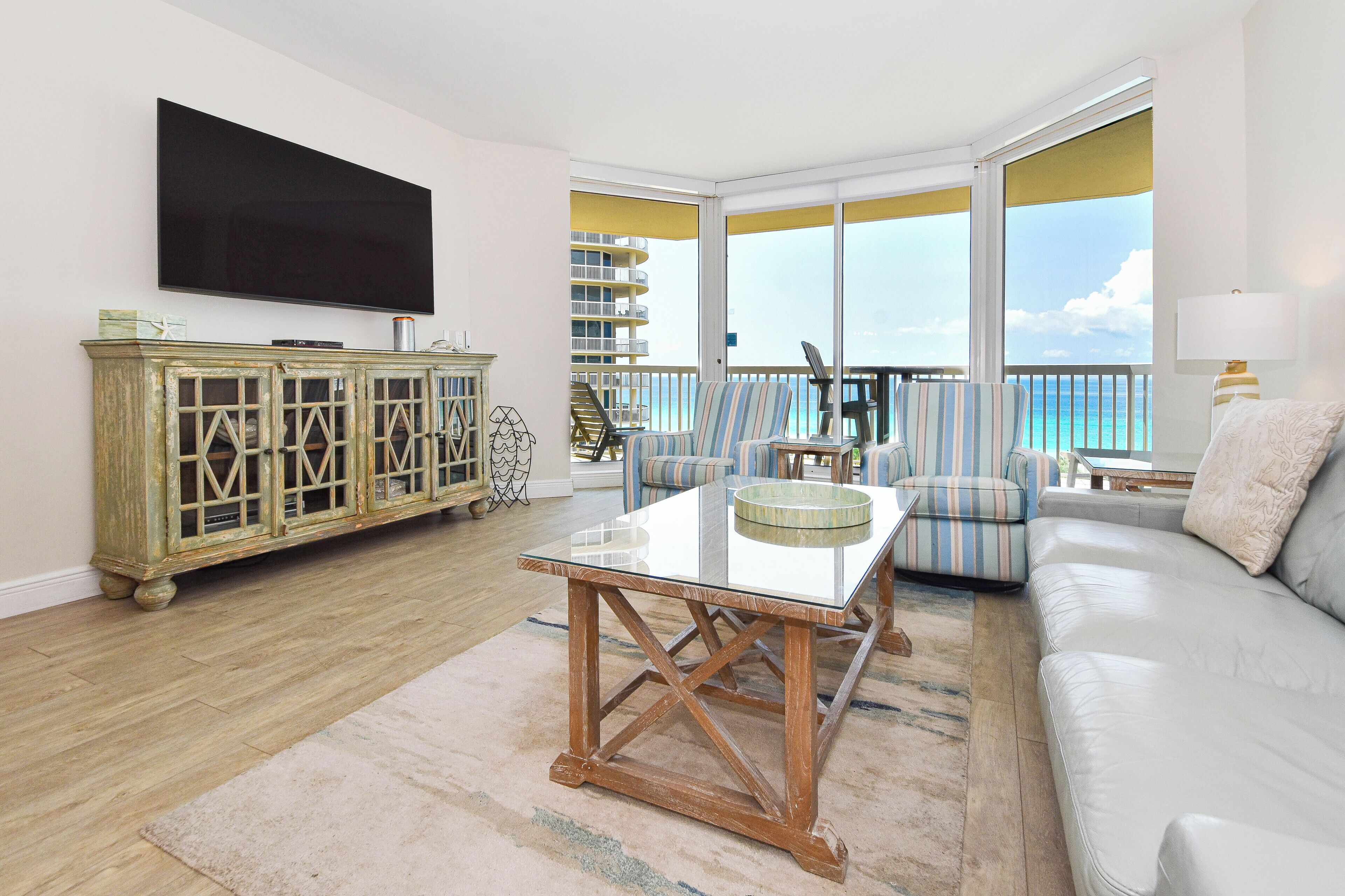 A living room with a flat-screen TV, a glass-top coffee table, a sofa, striped armchairs, and a view of the ocean through large windows.