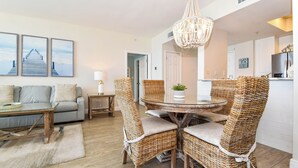 A living room with a sofa, a coffee table, and wicker chairs around a round table. There is a chandelier above the table. - Calm, cool, coastal beachfront condo w/private balcony! 🐚 (Destin)