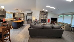 Une cuisine moderne avec un îlot central, un coin repas avec des chaises en bois, un canapé gris et une cheminée en pierre.