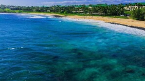 A beach with clear turquoise waters, a sandy shore, and a resort area with buildings in the background. - Ocean View & AC! 3 Min Walk To Poipu Beach! (Koloa)