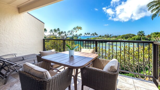 A balcony with wicker furniture, a table, and a view of a body of water and palm trees. - Ocean View & AC! 3 Min Walk To Poipu Beach! (Koloa)