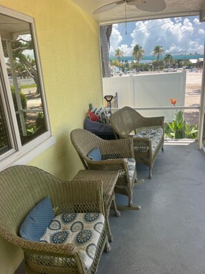 A covered outdoor seating area with wicker furniture and cushions, a view of the street and palm trees, and a ceiling fan. - Location!  Welcome To Bare Feet Retreat.  Steps From Times Square And The Beach (Fort Myers Beach)