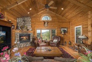 A log cabin interior with a stone fireplace, leather armchairs, a wooden coffee table, and a ceiling fan.