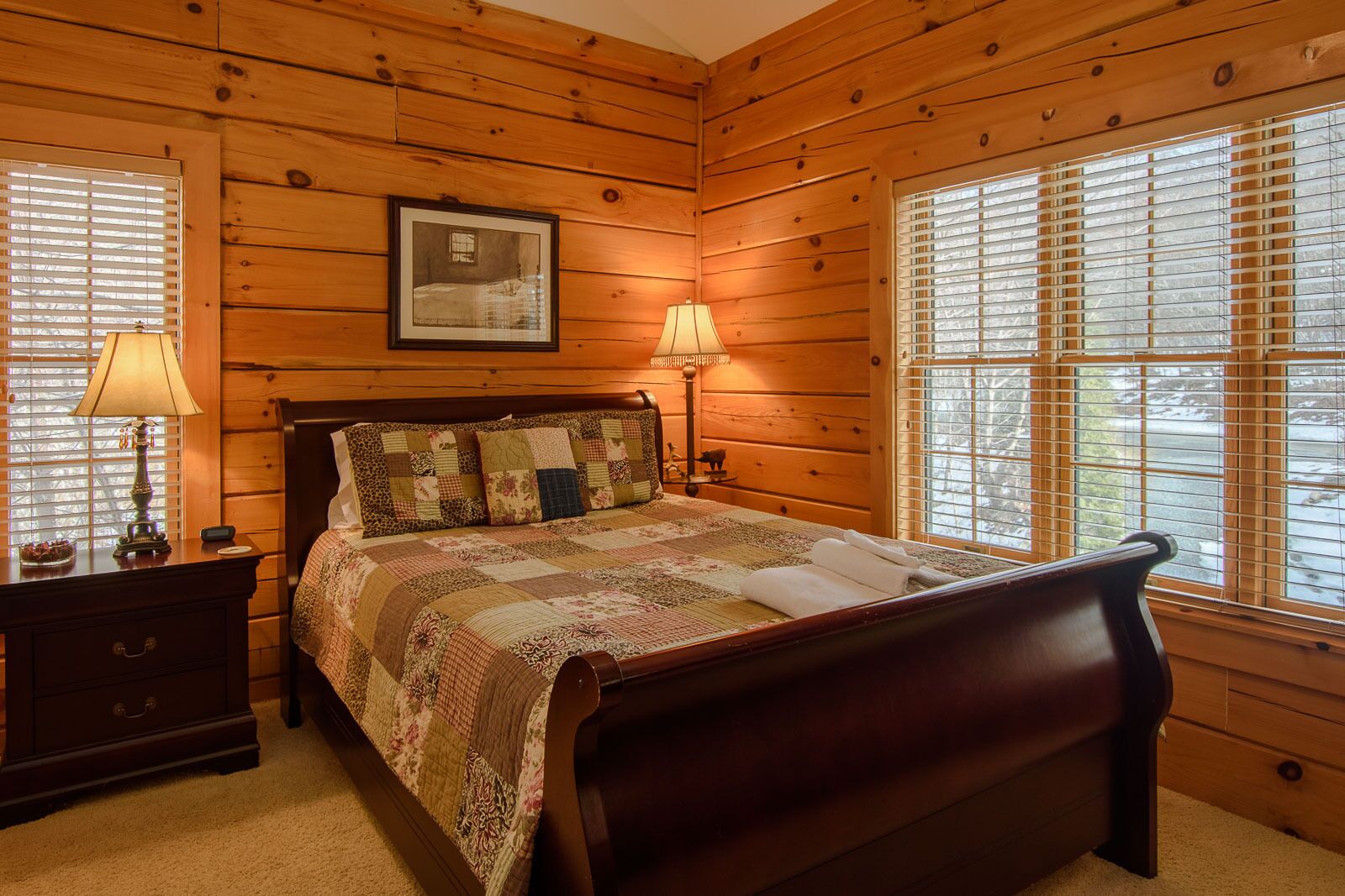 A wooden-paneled bedroom with a bed, two bedside lamps, a nightstand, and a framed picture on the wall.