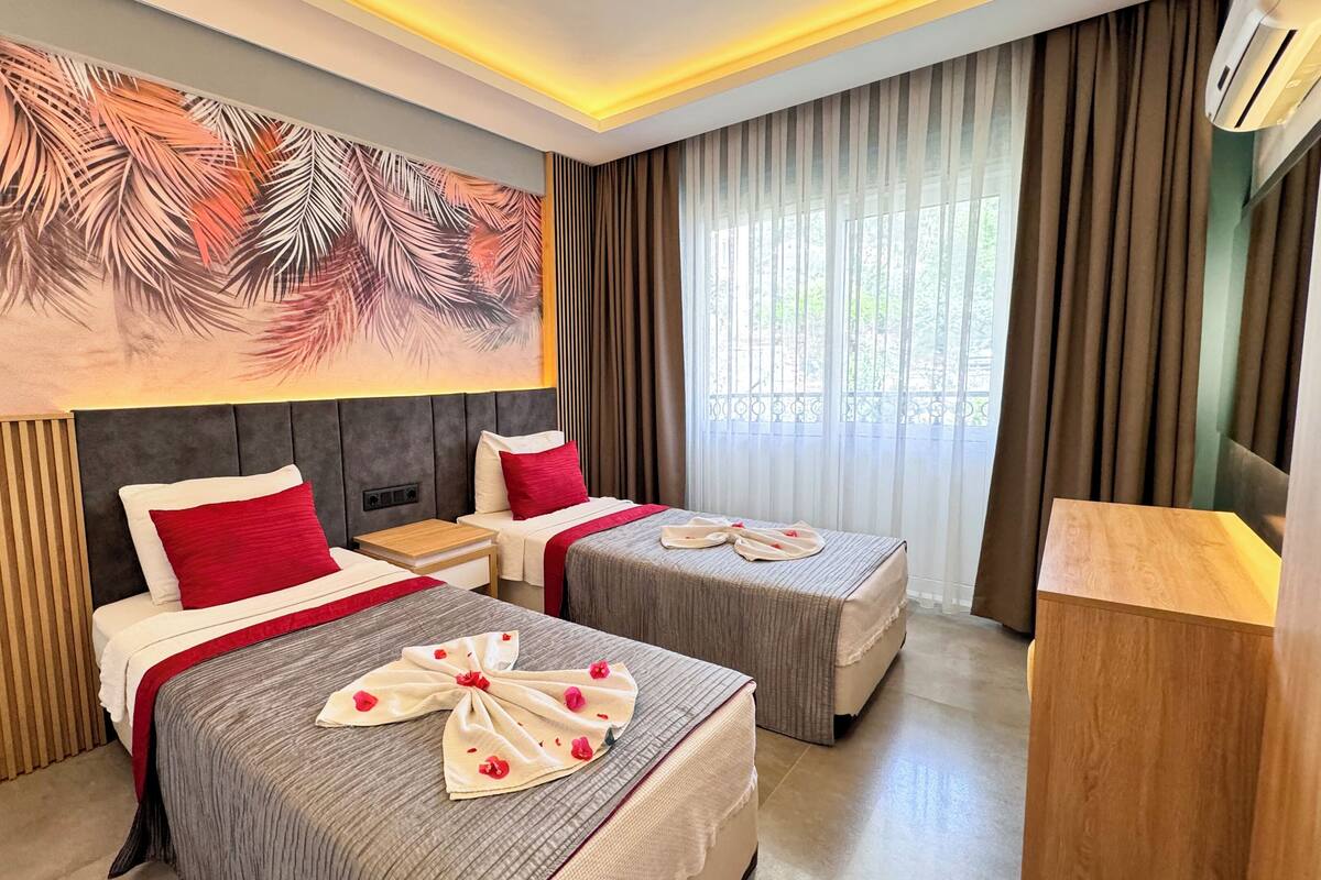A hotel room with two beds, a large tropical leaf mural, a wooden nightstand, and a large window with curtains.