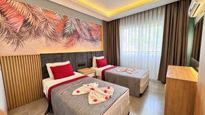 A hotel room with two beds, a large tropical leaf mural, a wooden nightstand, and a large window with curtains. - Gurol Hotel & Apart (Fethiye)