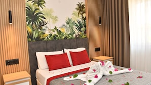 A hotel room with a bed, red pillows, a grey and white striped bedspread, a wooden nightstand, a wooden panel wall, a mural of tropical plants, and a curtain. - Gurol Hotel & Apart (Fethiye)