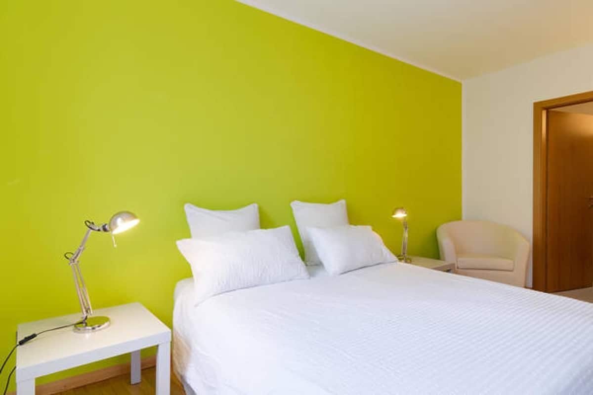 A bedroom with a bed, two bedside lamps, a chair, and a wooden door.