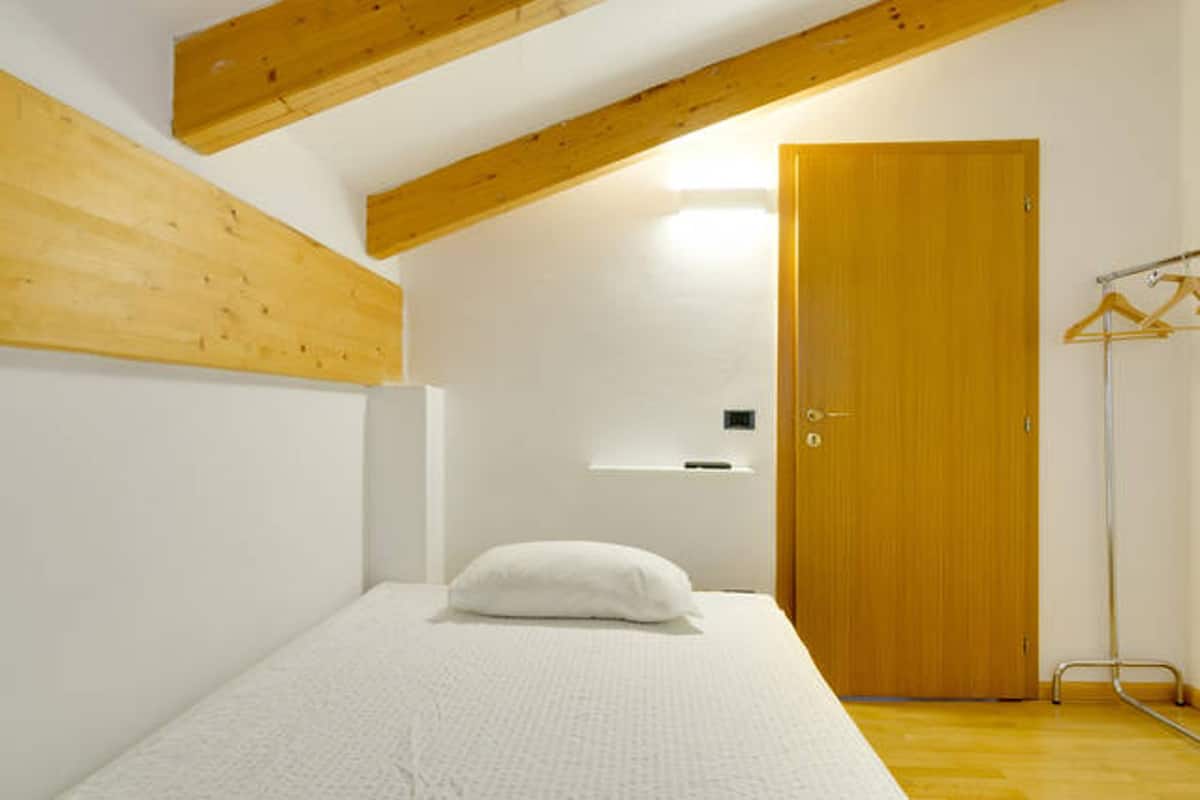 A bedroom with a wooden ceiling, a bed with a white mattress and pillow, a wooden door, and a coat rack.