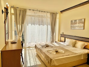 A hotel room with a bed, a bedside table, a nightstand, a window with curtains, and a painting on the wall. - Menada Andalusia Apartments (Elenite)