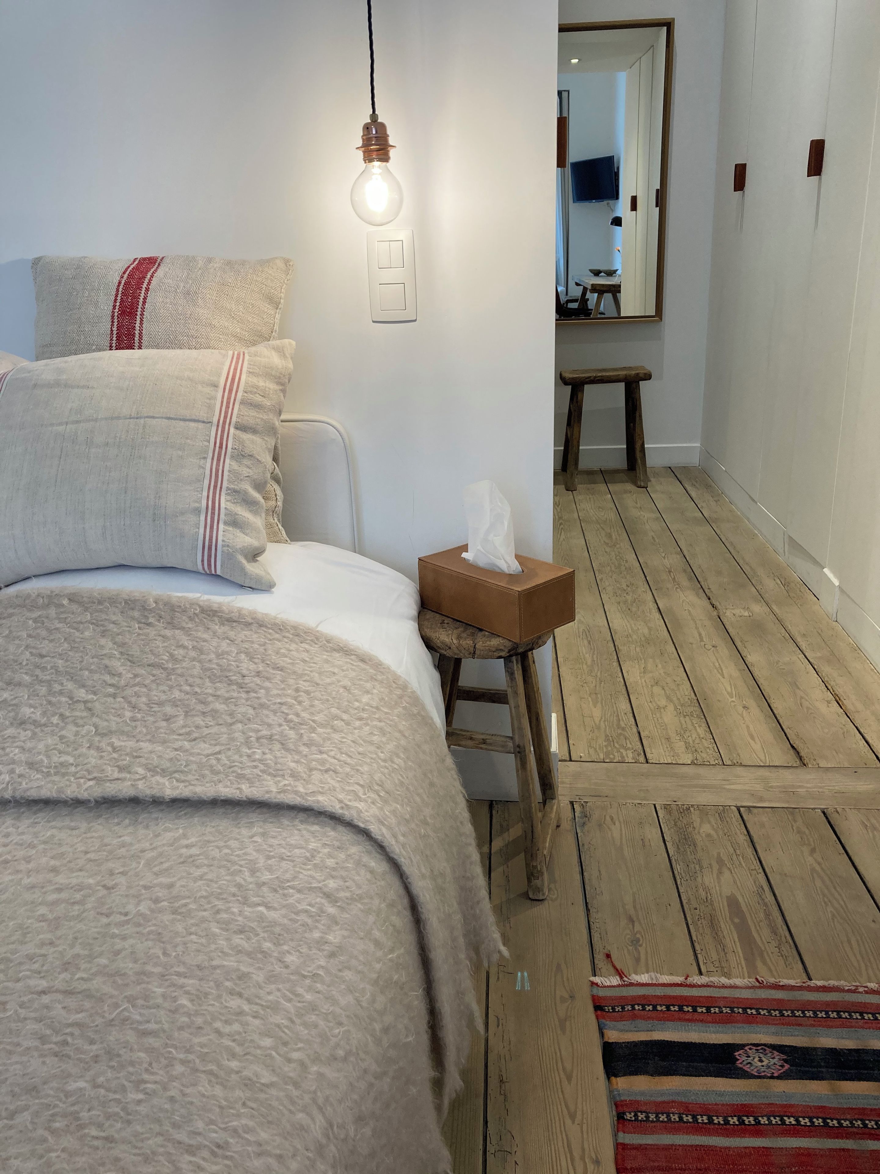 Un dormitorio con cama, una mesita de noche con caja de pañuelos, un taburete de madera, una lámpara y un espejo.
