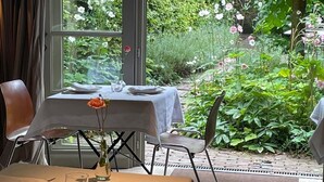 A dining area with a table set for two, a vase with a single flower, and a view of a lush garden through a window. - Hotel Hemelhuys (Hasselt)