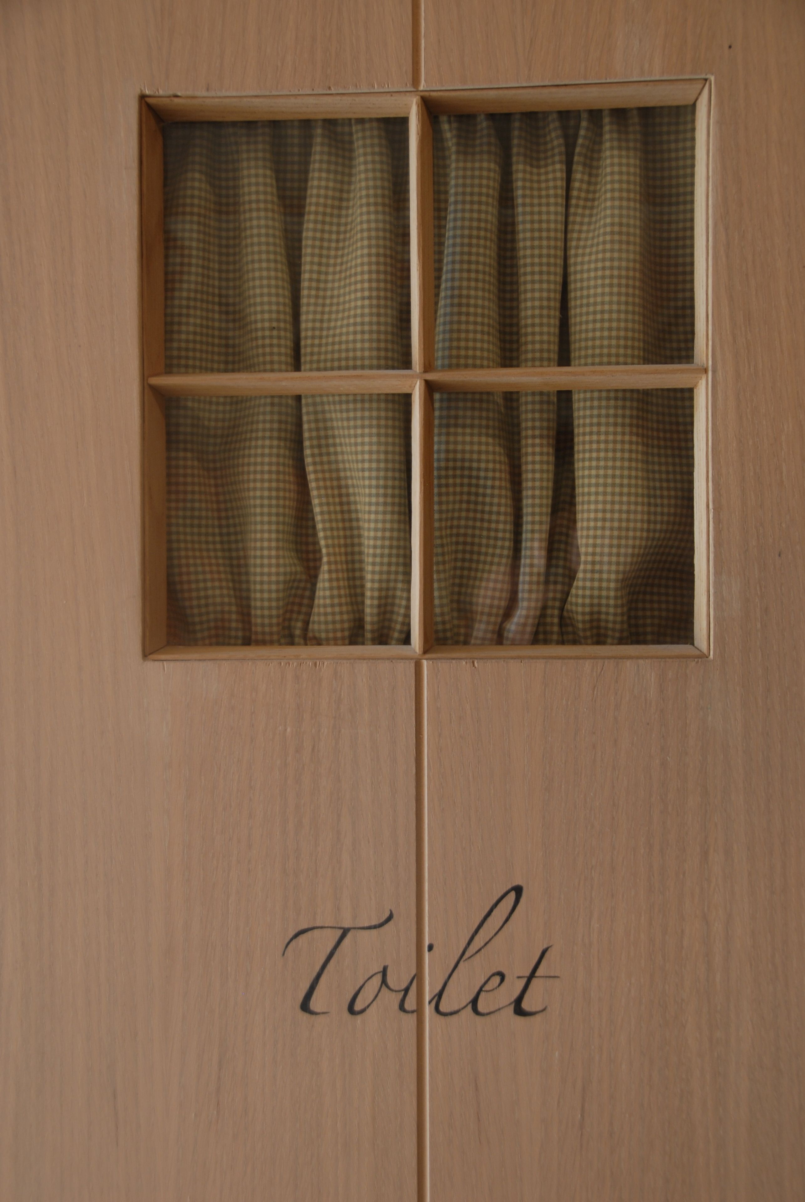 A wooden door with a window divided into six panes, each with a checkered curtain. The word "Toilet" is written below the window.