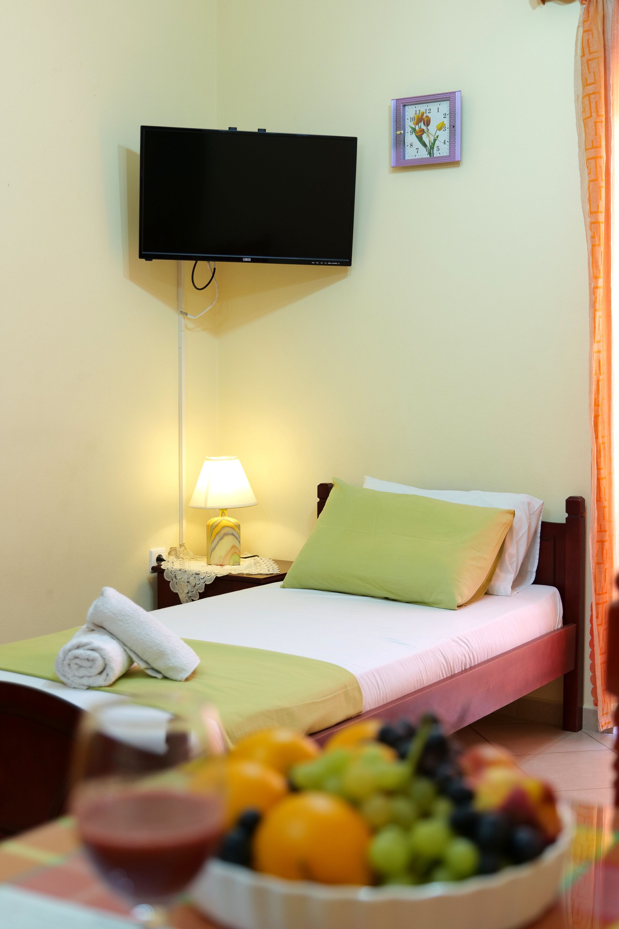 A hotel room with a bed, a bedside table with a lamp, a flat-screen TV mounted on the wall, a fruit bowl on the table, and a window with curtains.