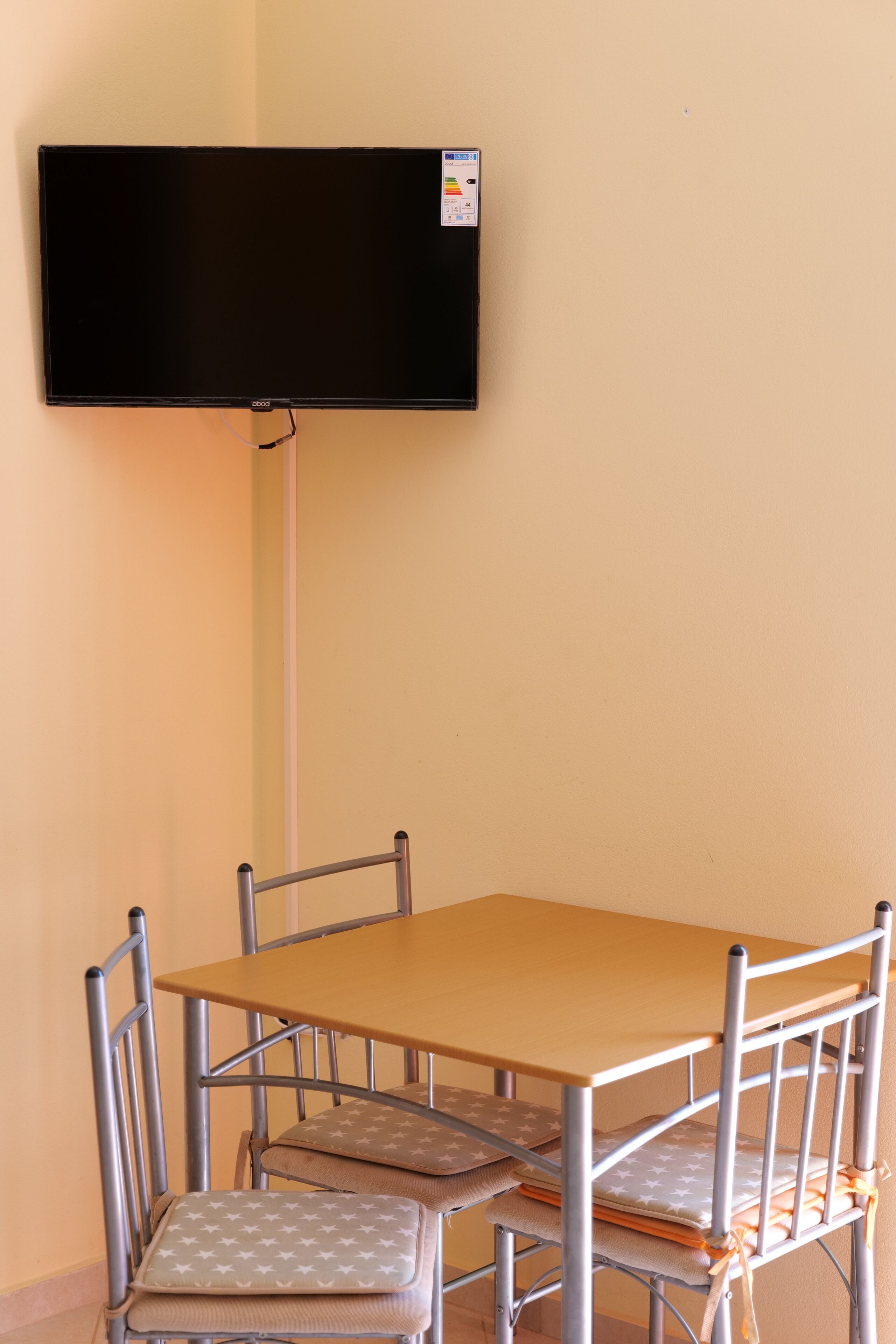 A dining area with a table and four chairs, a flat-screen TV mounted on the wall, and a wall-mounted electrical outlet.
