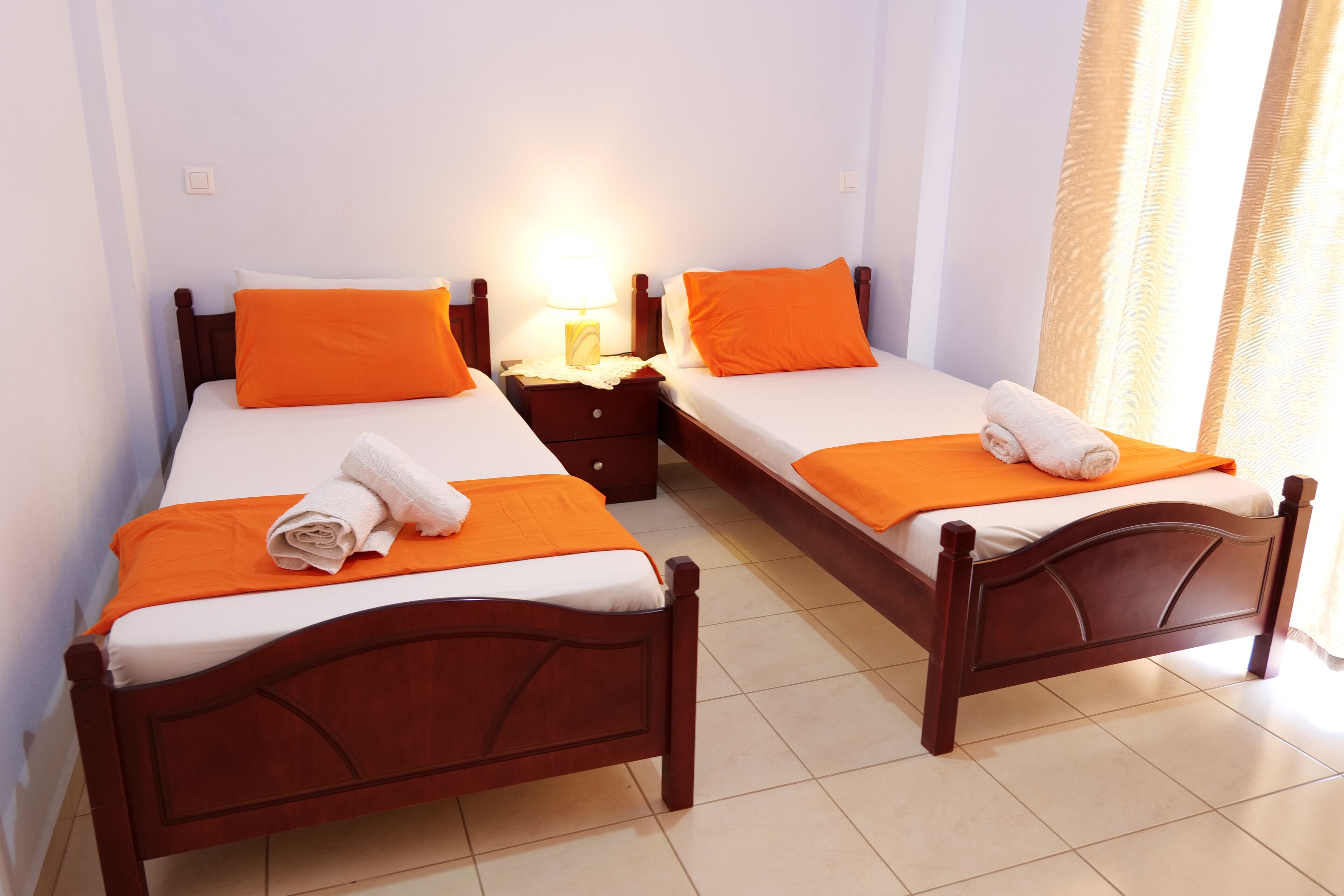 Two single beds with wooden frames, white bedding, and orange pillows. Each bed has a small bedside table with a lamp. The room has white walls and a tiled floor.