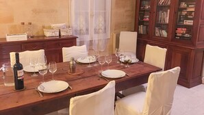 A dining table set for six with white chairs, a wine bottle, and glasses. A cabinet with books and board games is in the background. - Les Gués Rivières (Pujols)