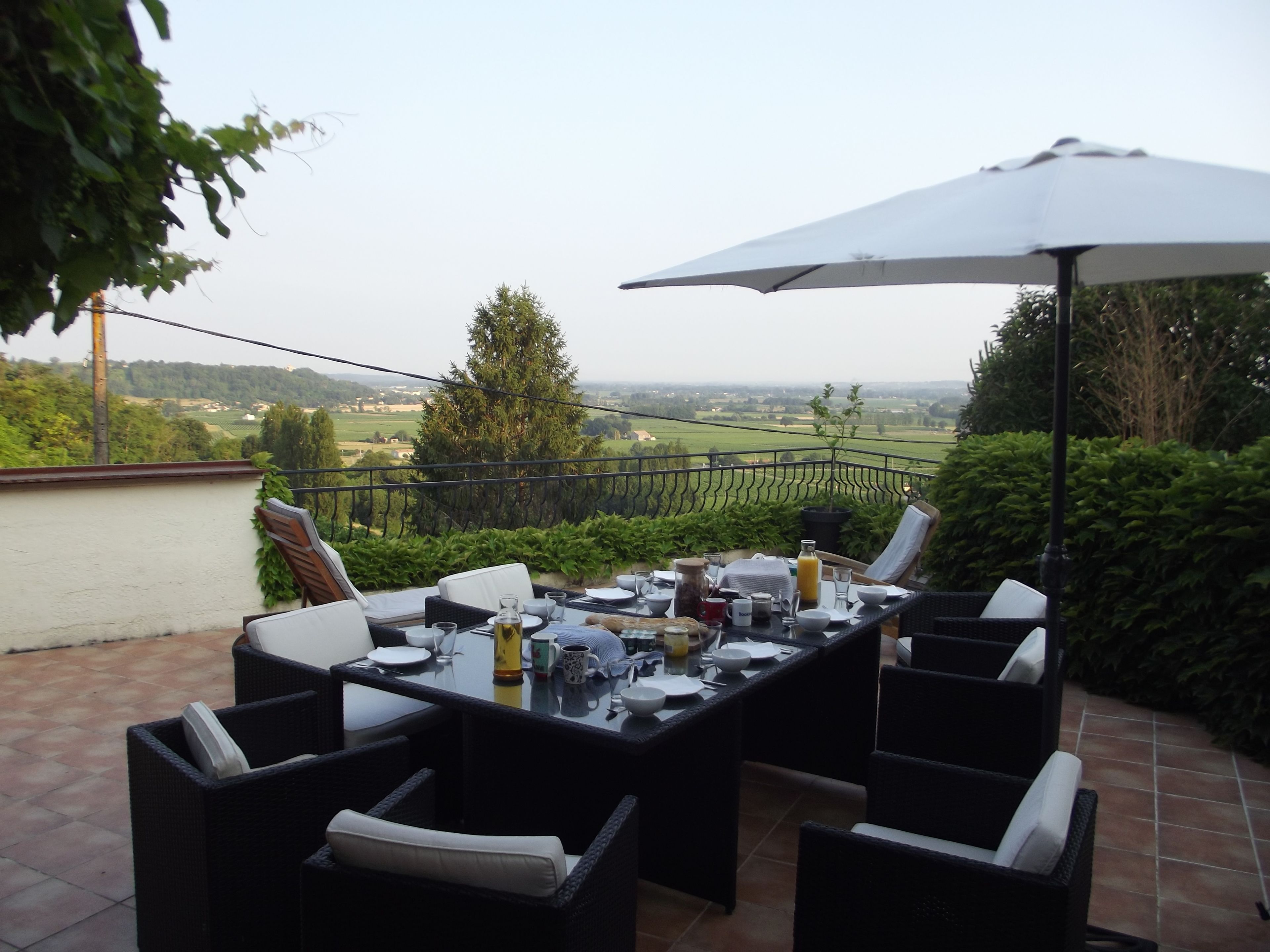 An outdoor patio with a table set for dining, surrounded by wicker chairs, and an umbrella providing shade.