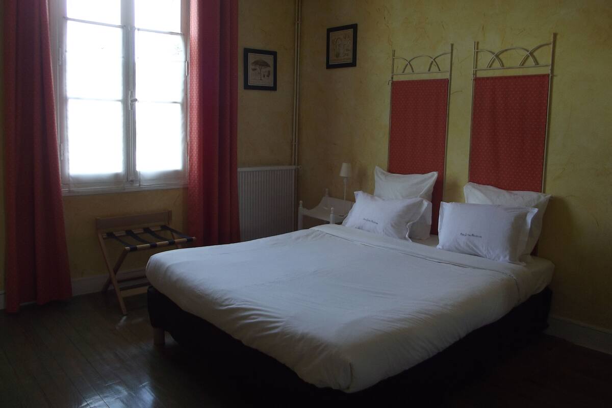 A hotel room with a bed, a radiator, a window with curtains, and a wall-mounted shelf.