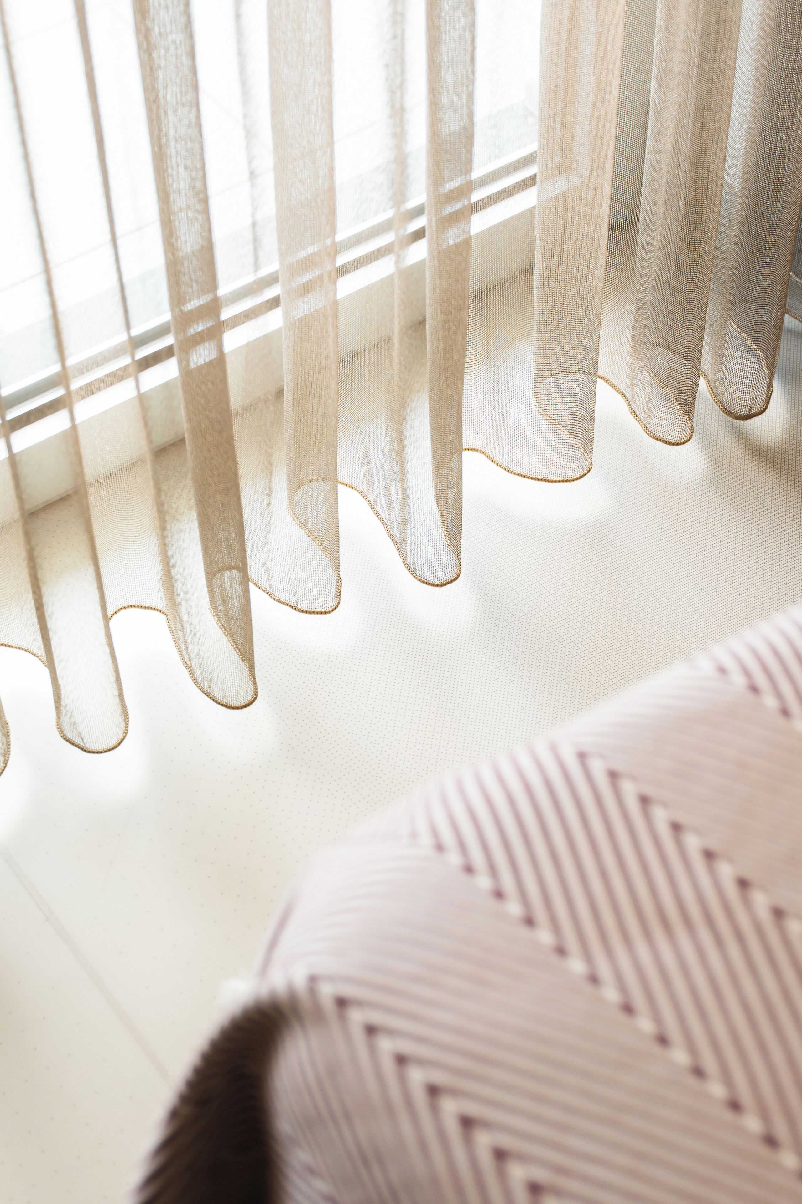 Primer plano de cortinas translúcidas con borde ondulado, un cojín estampado y un suelo de tonos claros.