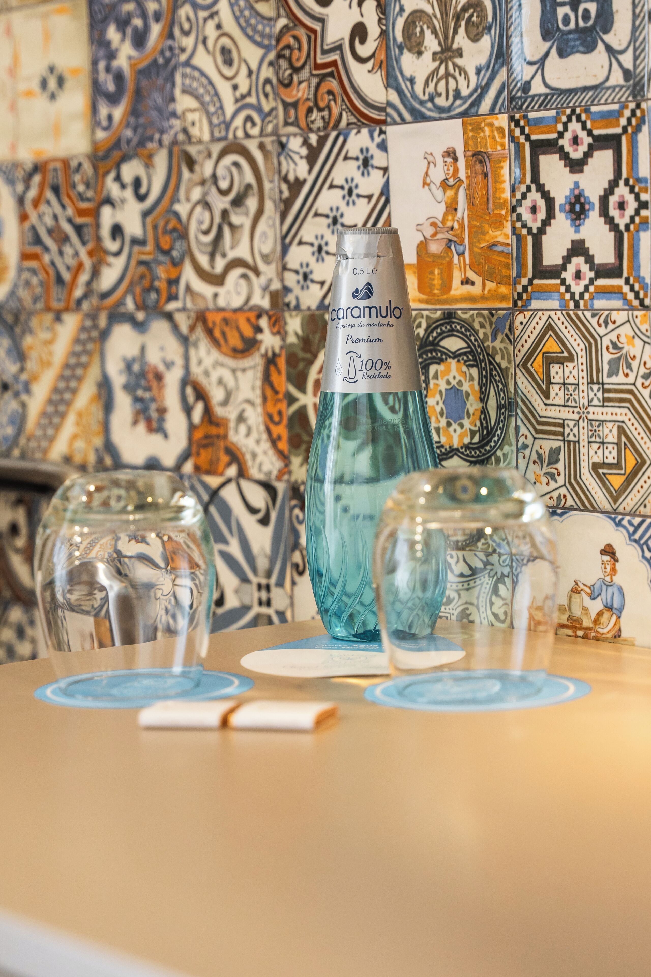 A table with a bottle of caramulo premium water, two clear glass vases, and a white candle. The background features a wall with colorful, patterned tiles.