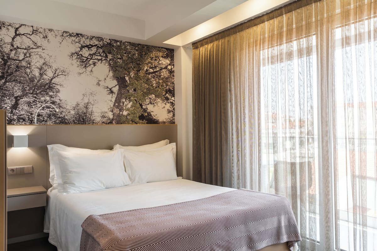 A hotel room with a large bed, a nightstand, a wall-mounted lamp, and a window with sheer curtains.