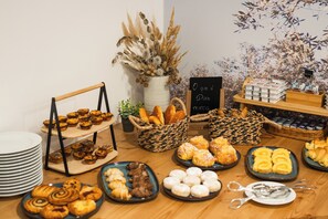 Una mesa de bufé con diversos pasteles y panes, incluyendo cruasanes, muffins y bollos. Hay cestas y platos para servir, y un cartel que indica que la comida es sin gluten.
